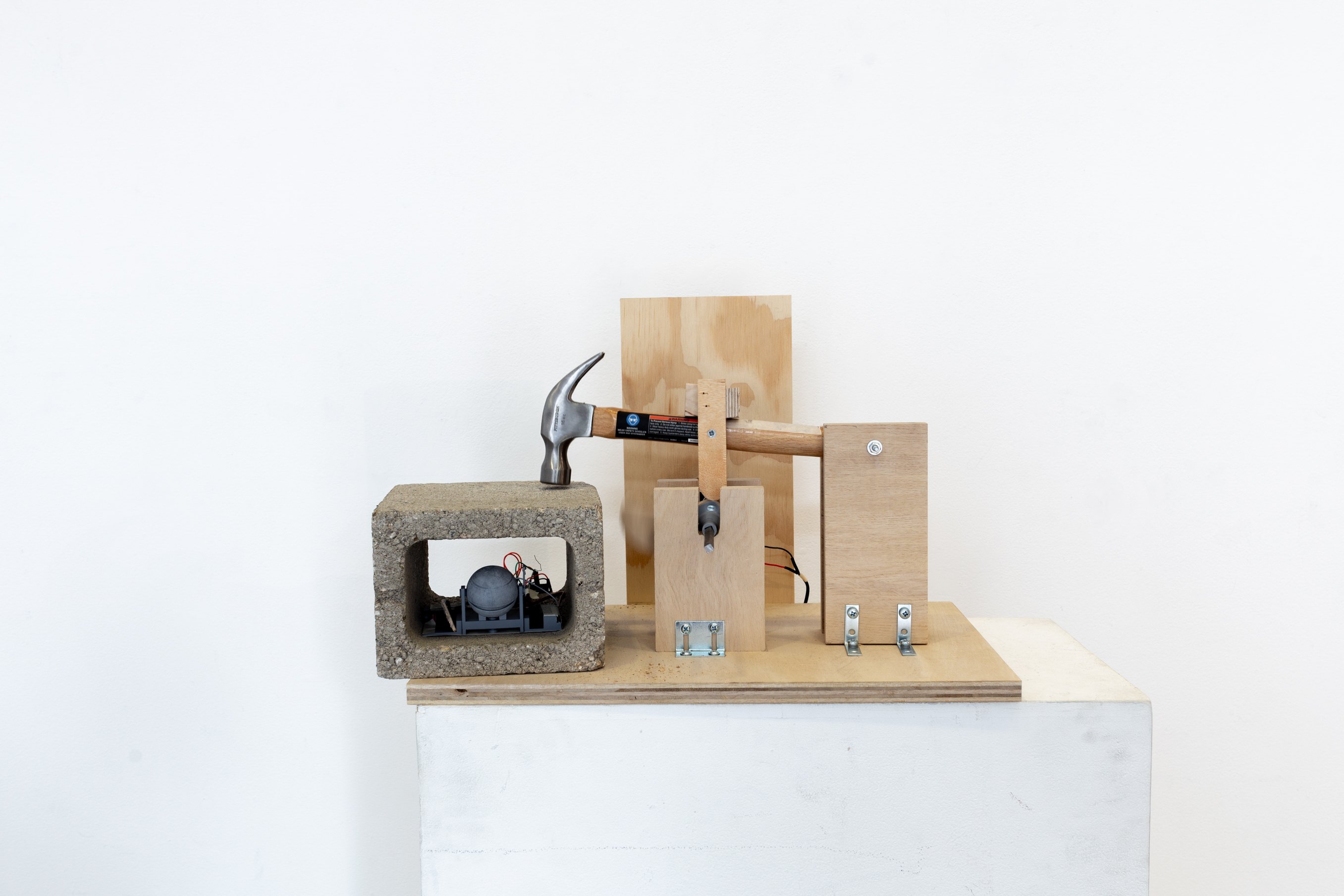 A sculpture consisting of a hammer assembeled by wood with electronics, where the hammer strikes a cinderblock, against a white wall.